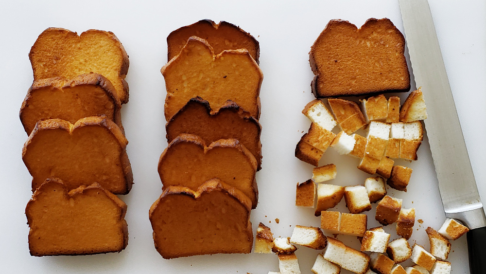 10 pieces of toasted angel food cake lined up with 1 piece cut into cubes on a white board