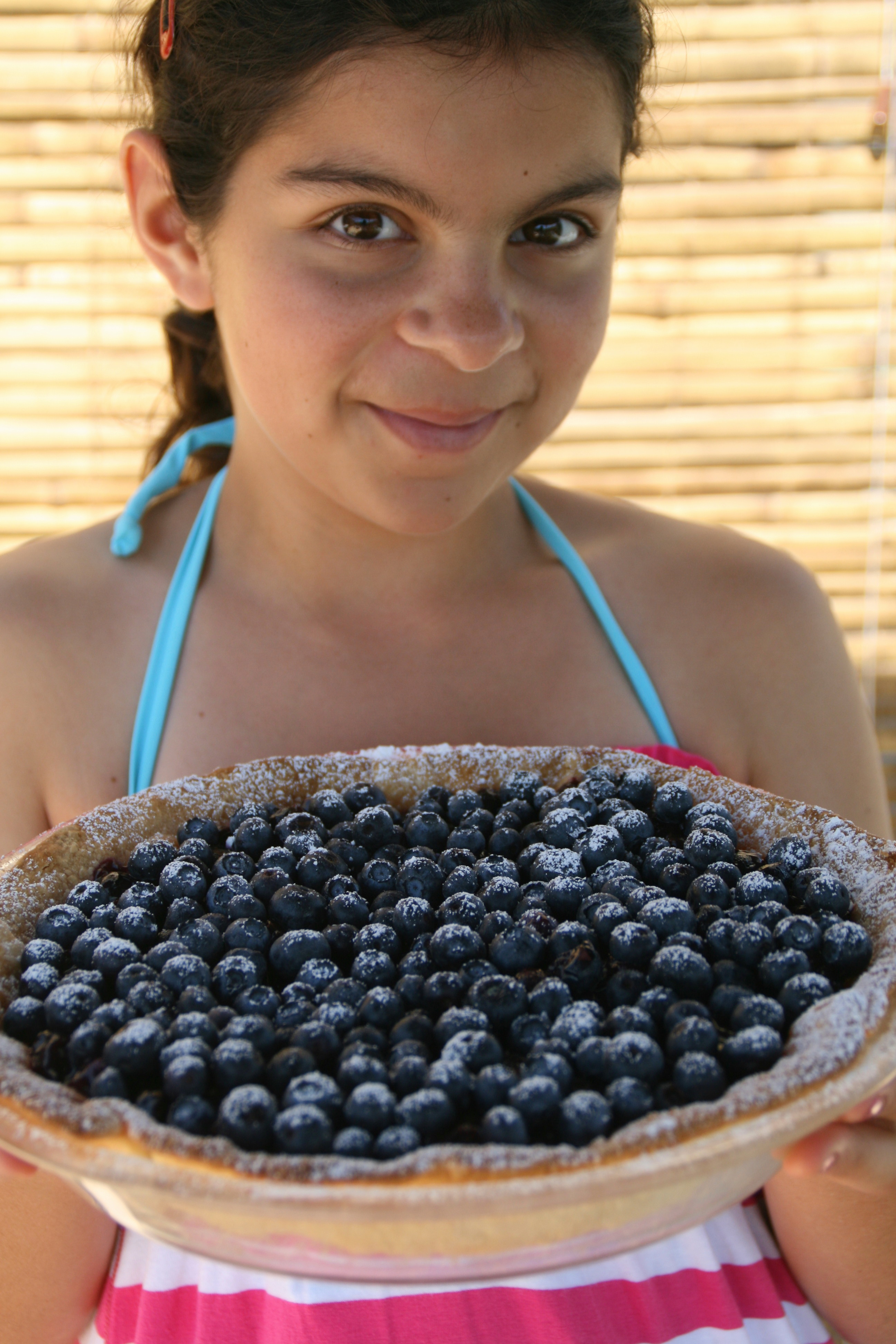 Young girl holds a Fresh Blueberry Pie sprinkled with powdered sugar against a bambook shade backdrop