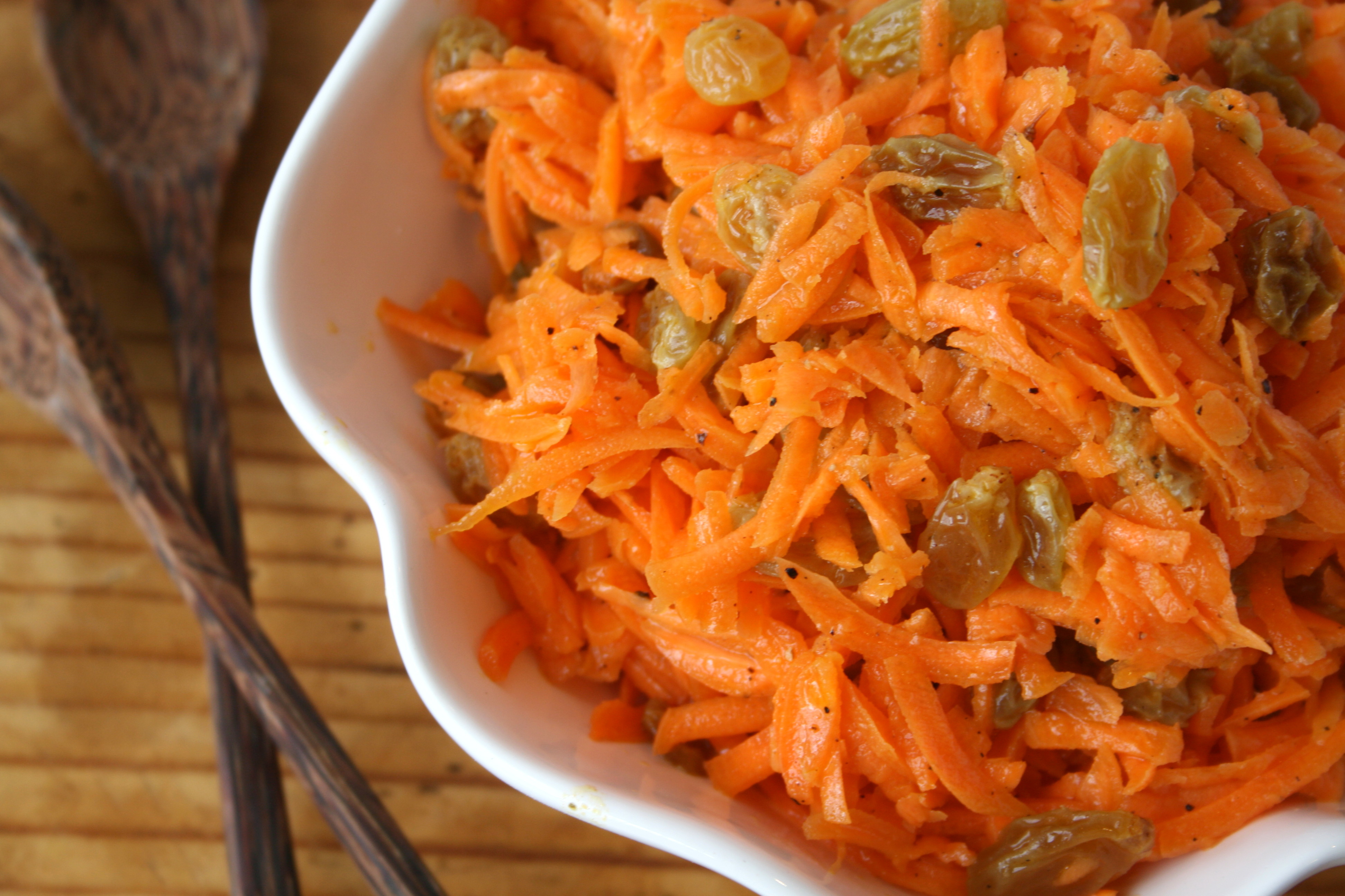 Bright orange carrot and raisin salad in a white bowl with wooden spoons along the left side