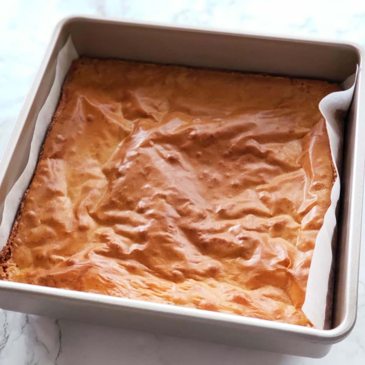 Blondies with a light brown rough surface are in a parchment-lined pan on a white marble counter