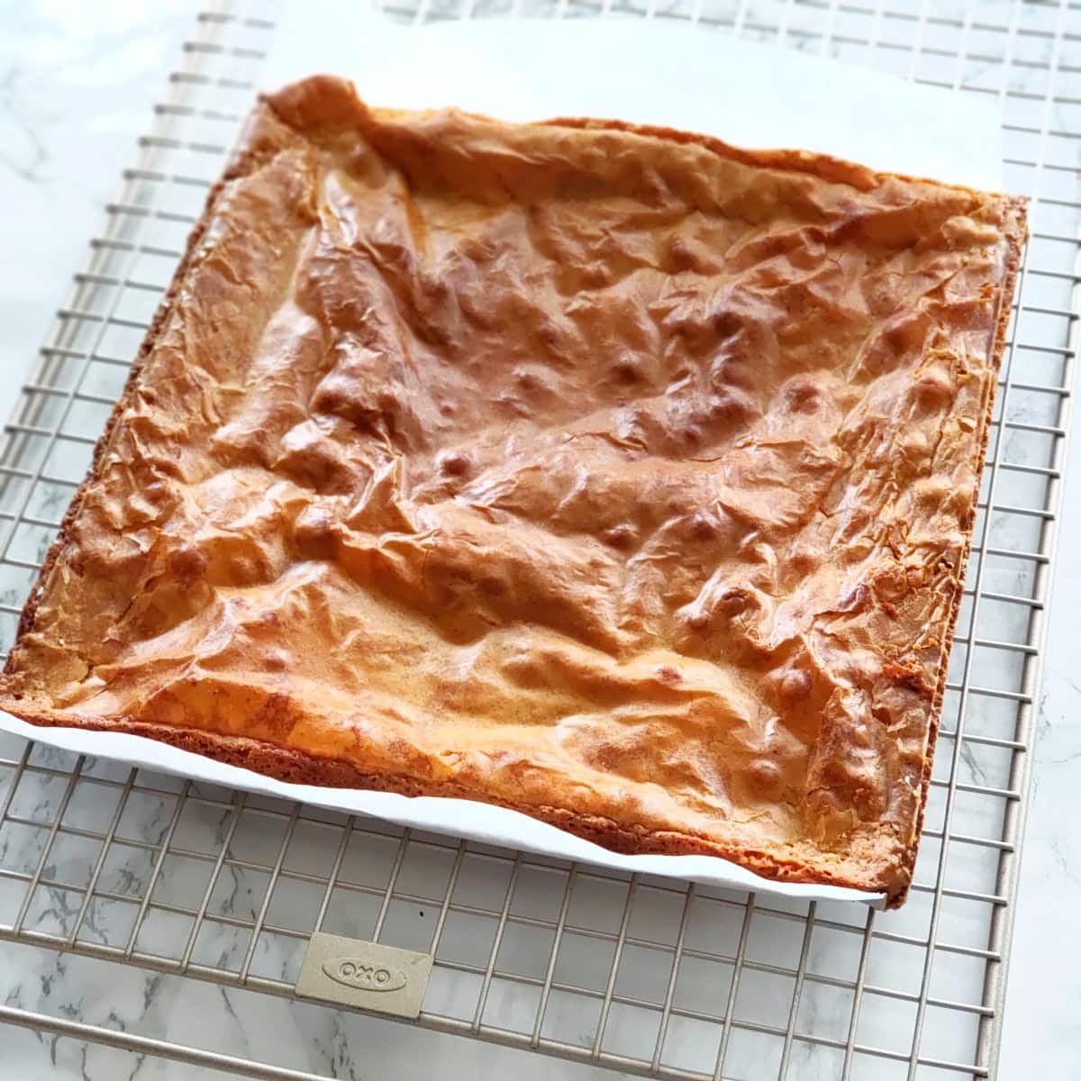 Square baked blondies with a light brown surface sit on their parchment paper on a cooling rack