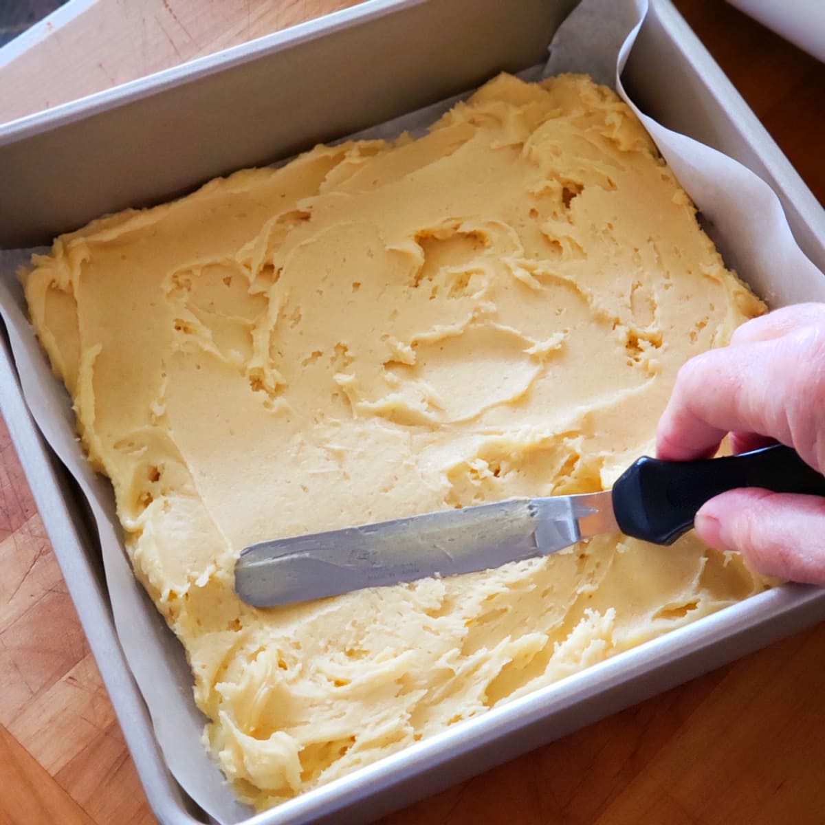 A hand holding an offset spatula smooths blondie dough into a parchment-lined baking dish