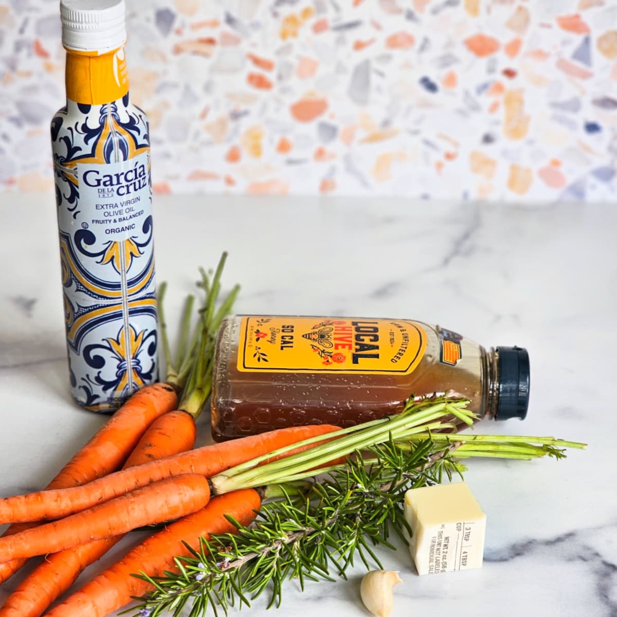 Ingredients for Honeyed Carrots on a white marble counter with a pattered wall behind. 