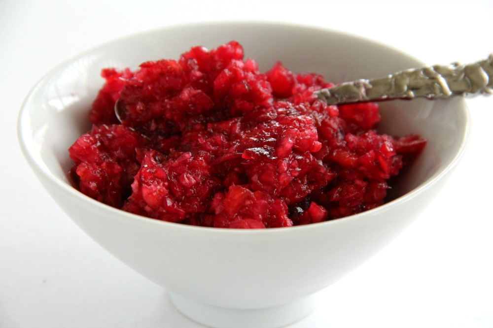 Bright red Fresh Cranberry-Ginger Relish in a white bowl with a spoon embedded against a white background