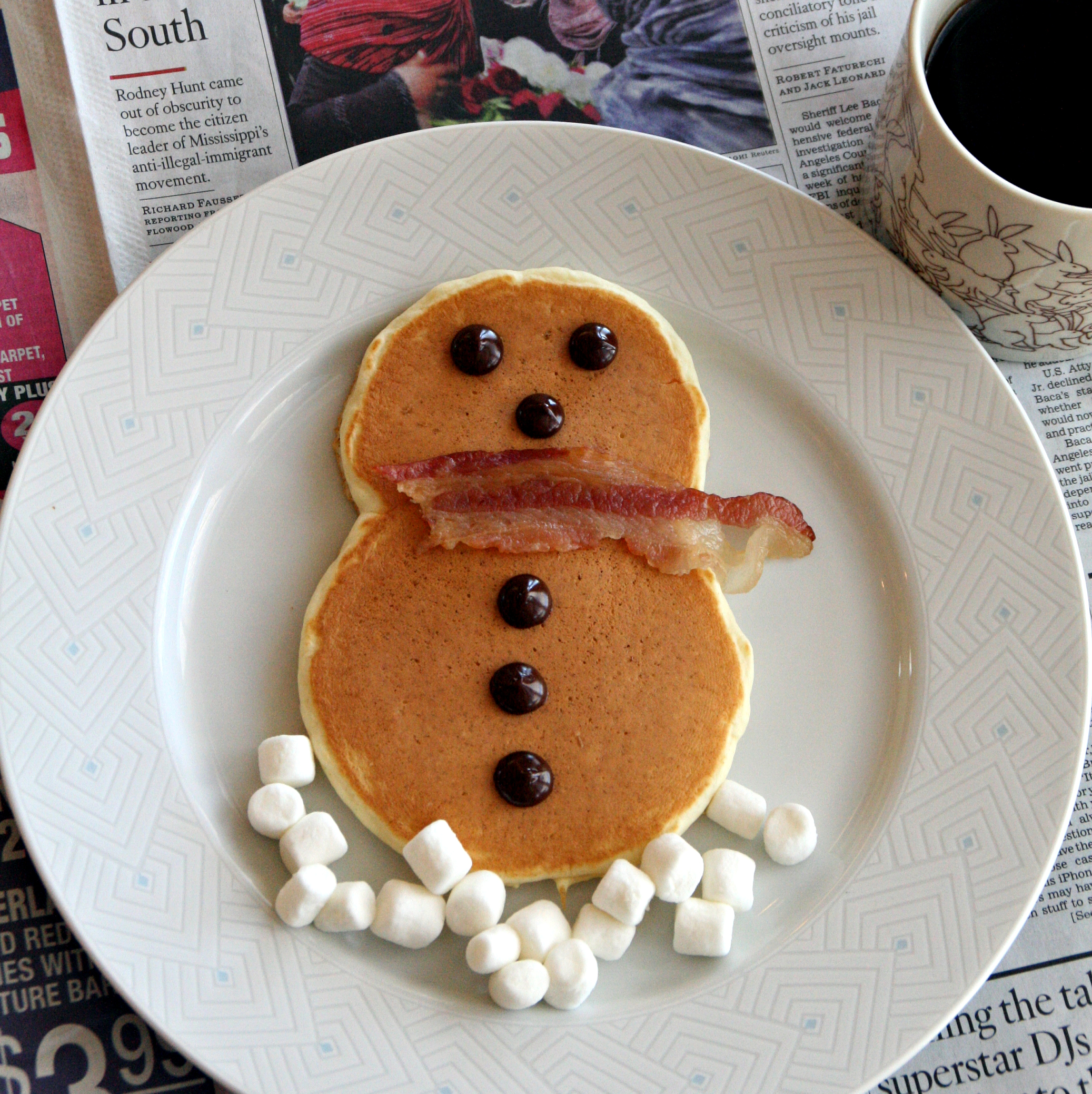 Pancake shaped like a snowman, with bacon as a scarf and marshmallows as the snow beneath and chocolate chips as the eyes and buttons