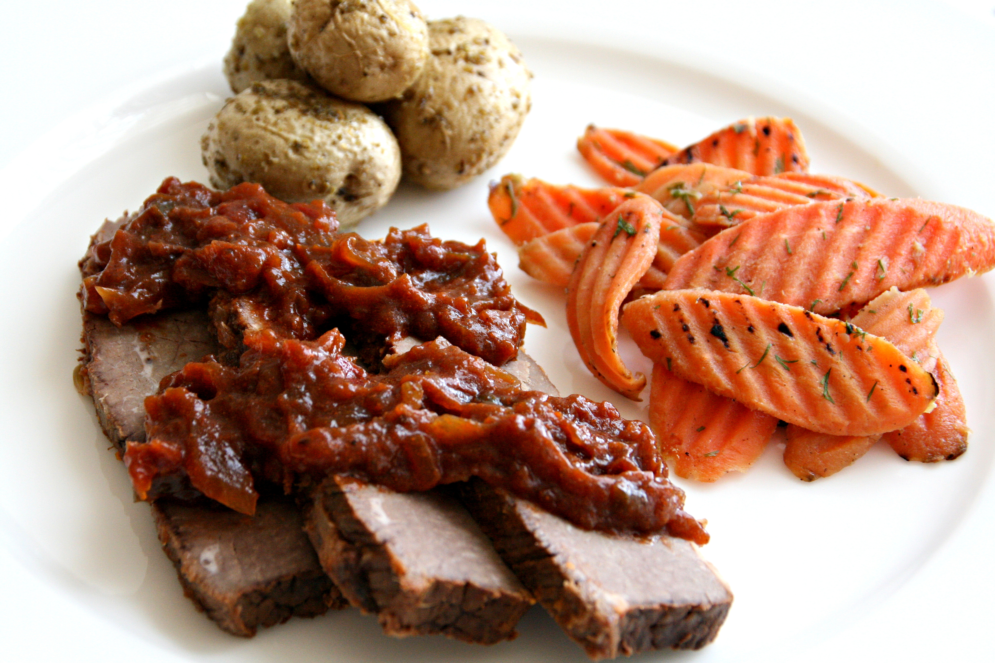 3 slices of brisket topped with red sauce, on a white plate, along with small potatoes and sliced carrots on ShockinglyDelicious.com