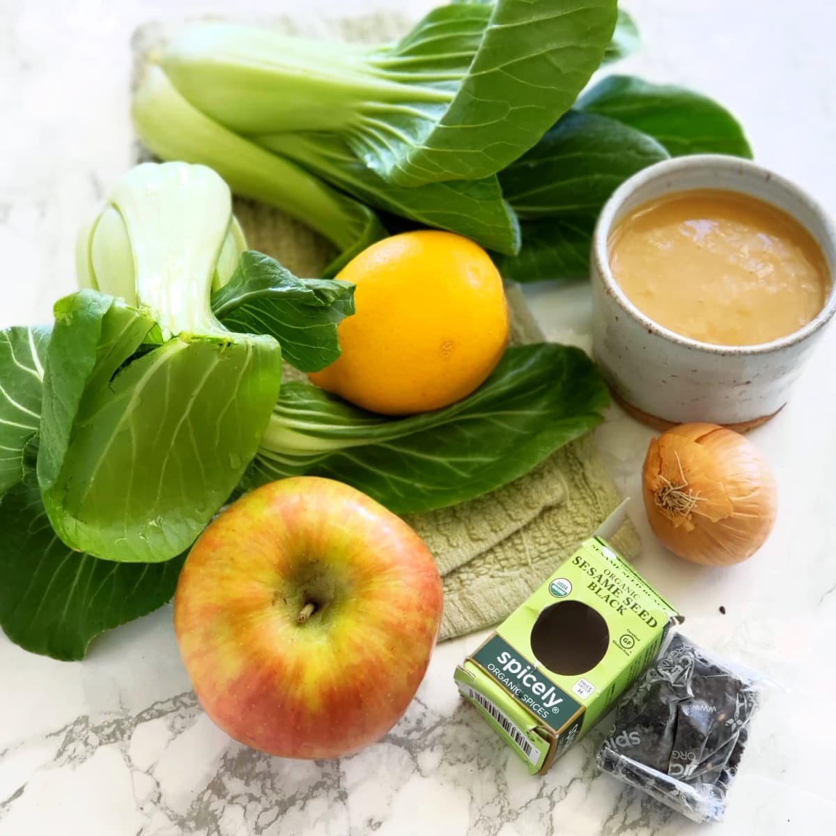 Ingredients for Lemony Baby Bok Choy Apple and Miso Salad -- bok choy, apple, lemon, shallot, black sesame seeds and dressing, on a marble countertop