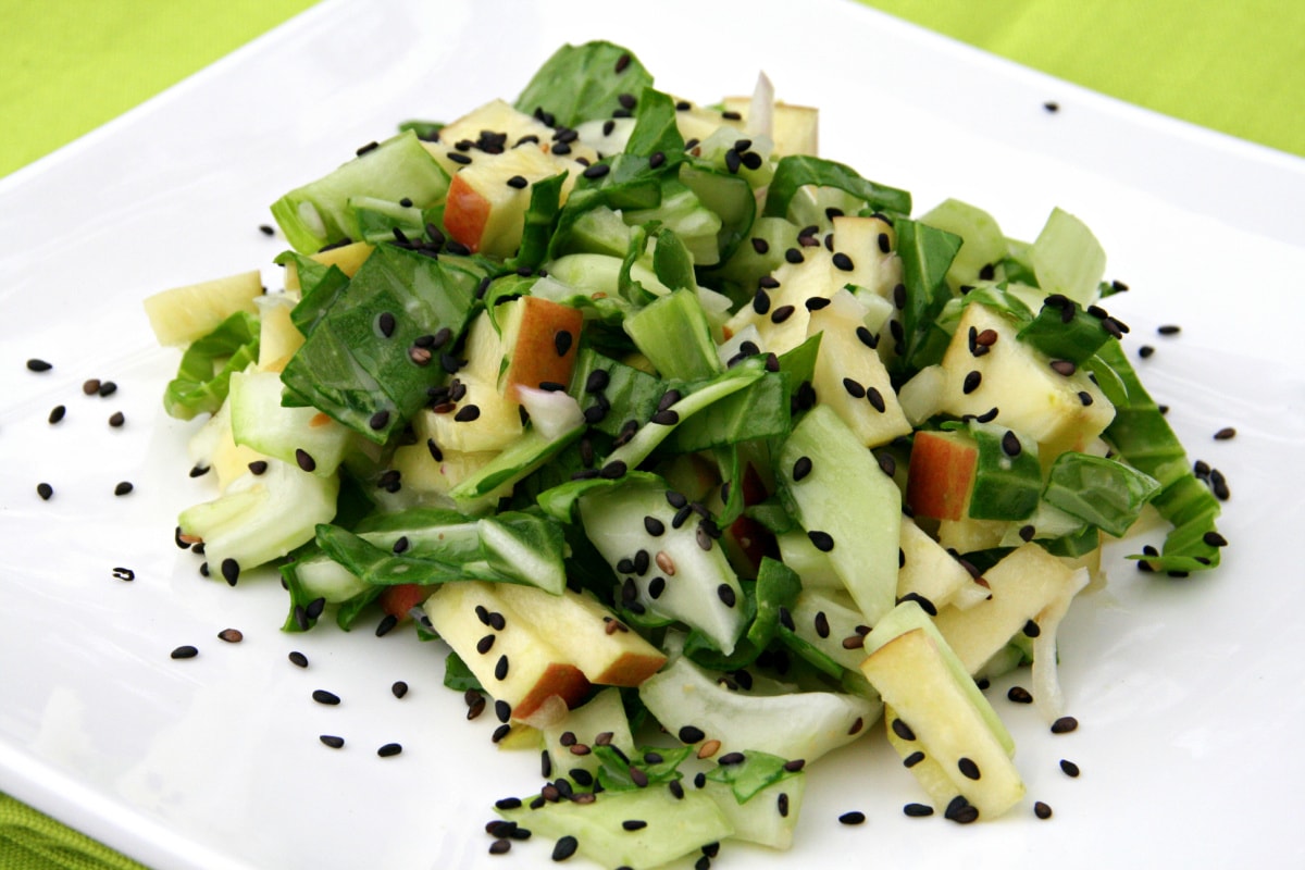 Baby bok choy and apple salad with lots of black sesame seeds sprinkled on top on a square white plate on a bright green placemat