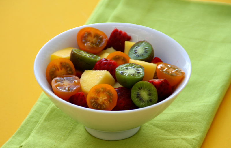 Fruit Salad with Tomatoes in a white bowl on a green napkin atop a bright yellow surface on ShockinglyDelicious