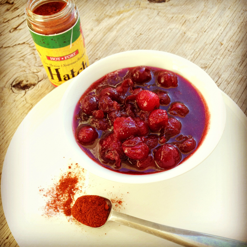 New Mexican Hatch Chile Cranberry Sauce in a white bowl on a white plate with a teaspoon of Hatch powder in the foreground and a bottle of Hatch powder in the background