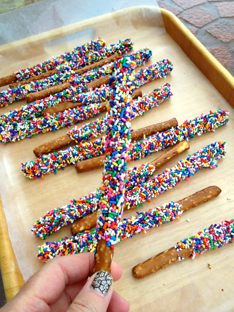 Decorated Festive Pretzel Rods are lined up on a gold tray, with a hand holding one to the camera. 