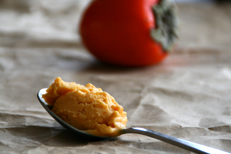 Persimmon Ice Cream on a spoon in front of a whole persimmon fruit on a wrinkled paper background