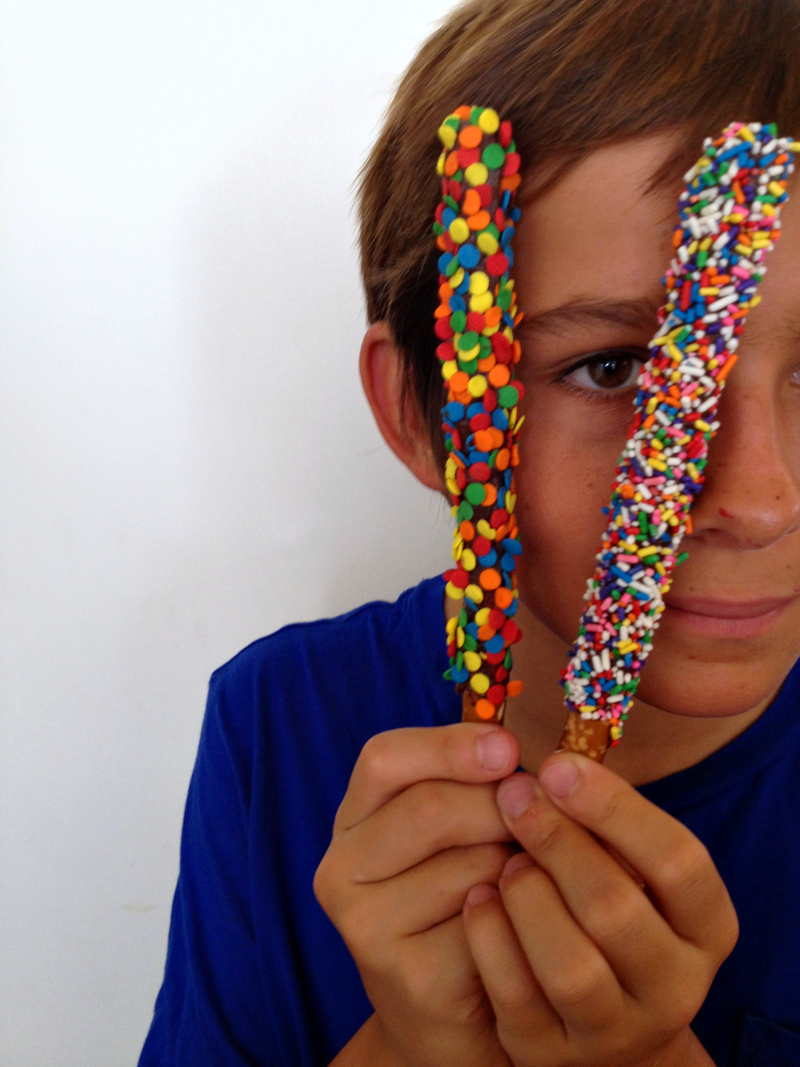 A boy in a blue shirt holds 2 decorated pretzel rods in front of his face and is peeking through them