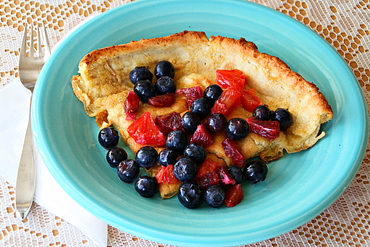 Dutch Baby Pancake on a bright turquoise plate on a cream colored lace tablecloth with a fork tucked under the left side of the plate