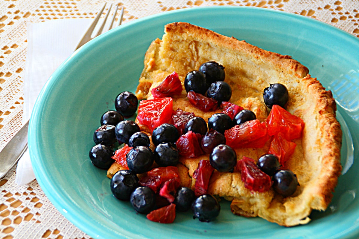 Dutch Baby Oven Pancake on a turquoise plate with blueberries and blood oranges on top, and a fork along the left side of the plate 