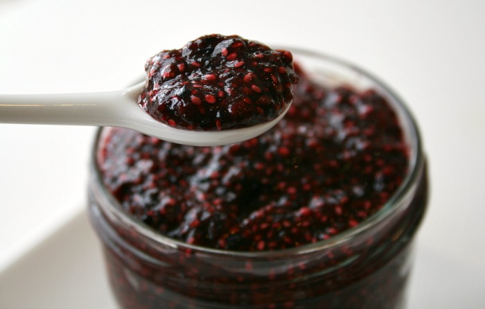 Dark purple jam with seeds in it on a white spoon above a jar of jam