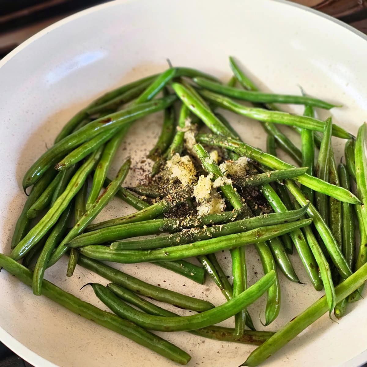 Garlic and herbs sit atop green beans in a cream-colored skillet 