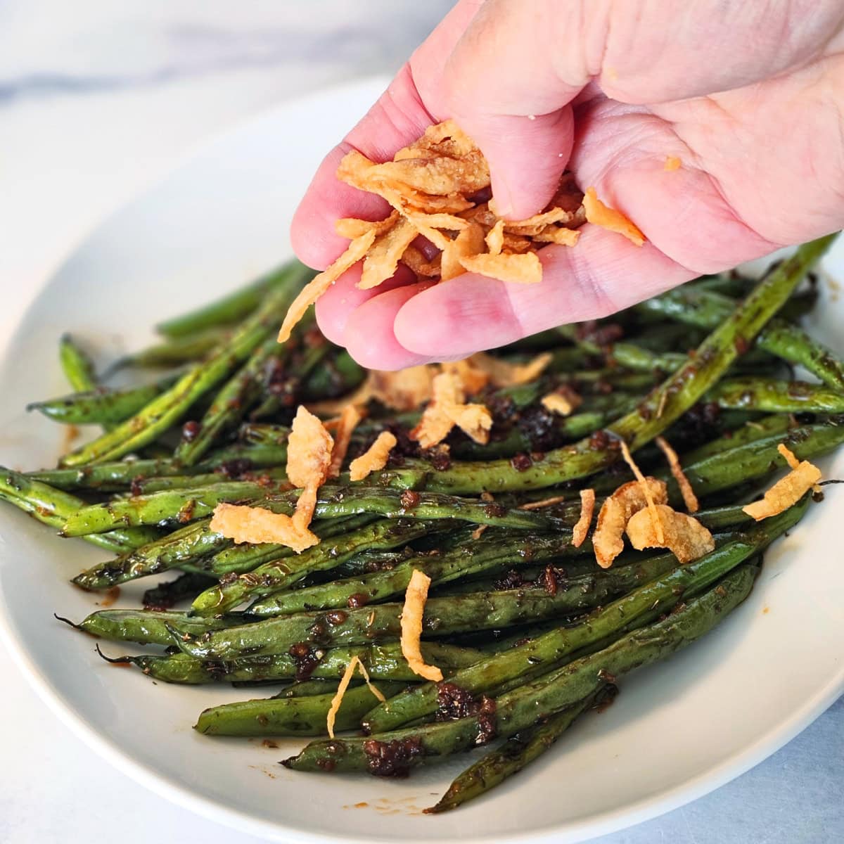 A hand sprinkles crispy fried onions on the Sweet and Savory Green Beans in a white bowl