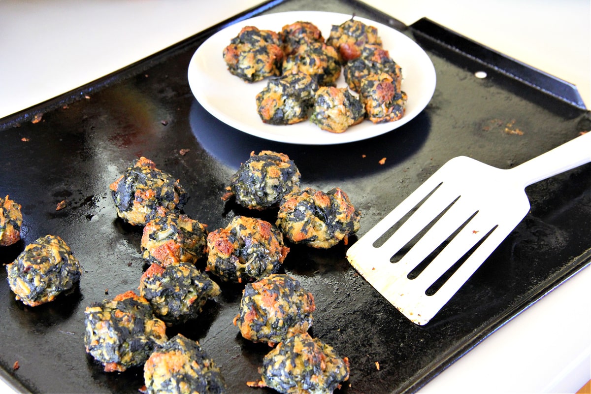 Spinach Balls on a black baking sheet with white spatula and white plate with a few balls on it