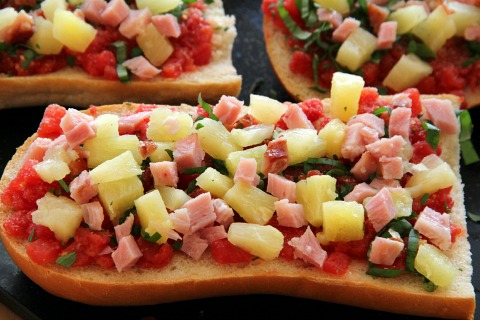 Assembling ingredients like ham, pineapple and tomatoes on top of French Bread Hawaiian Pizza 