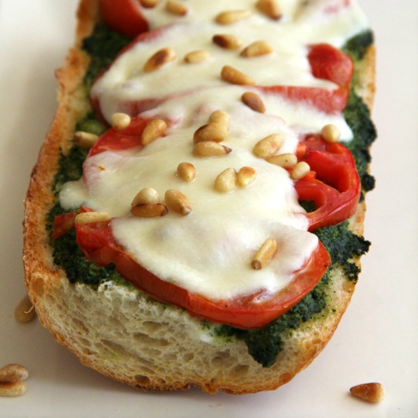 Half a loaf of French bread with pesto, tomato slices, melted Mozzarella and toasted pine nuts on top