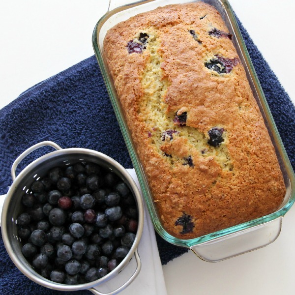 Loaf of blueberry bread resting on a purple towel with blueberries in a small colander alongside