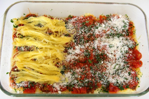 Yellow stringy spaghetti squash layered in a glass rectangular dish with tomato sauce and herbs and parmesan cheese on topyDelicious.com