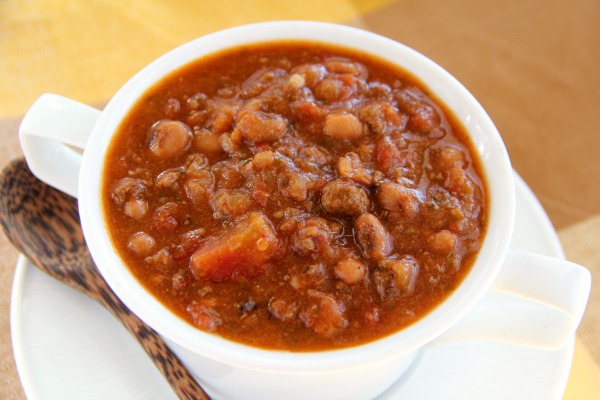 Brown colored Black-Eyed Pea Taco Soup in a white bowl on a yellow plaid tablecloth with a wooden spoon alongside