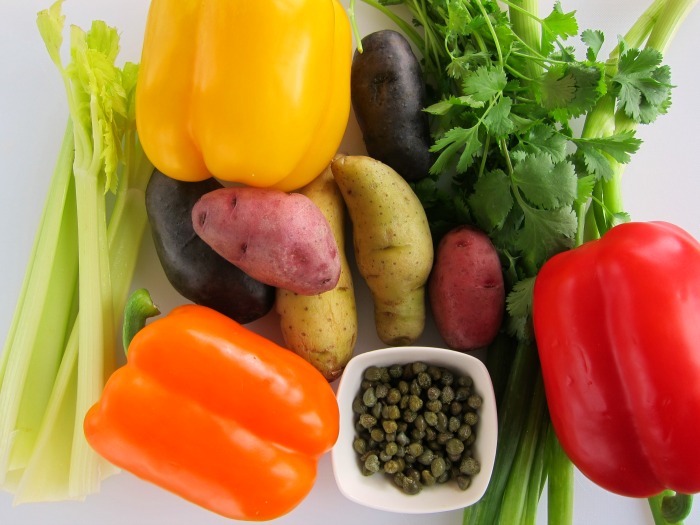 Ingredients for Grilled Rainbow Potato Salad on a white board -- celery, potatoes, bell peppers, cilantro, onion and capers