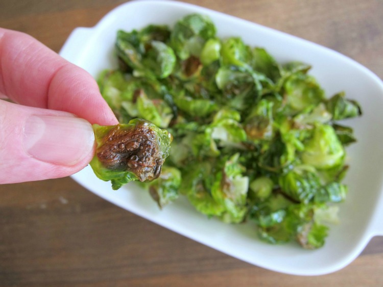 Fingers hold a single brussels sprout chips in the foreground, with a white plate of chips out of focus in the background