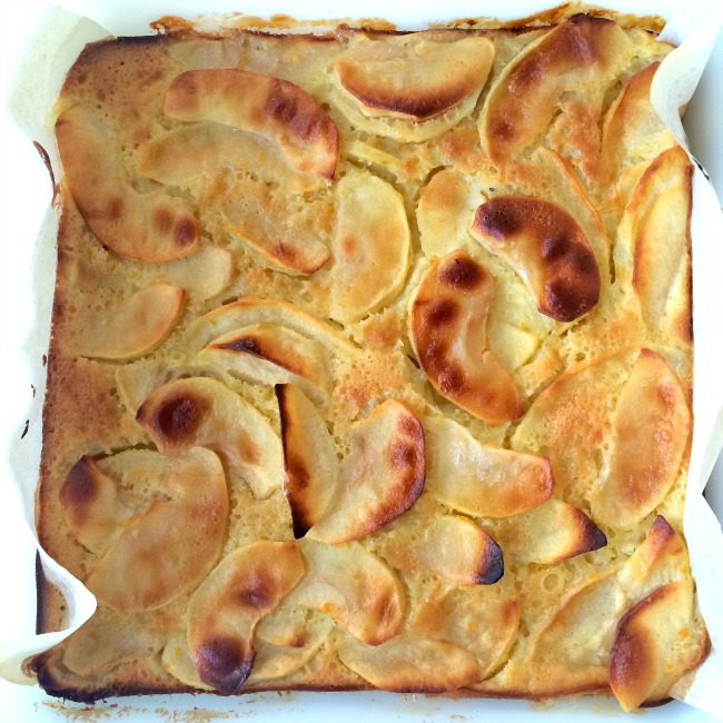 Browned Custardy Apple Squares in a square white baking dish with parchment paper peeking out from the sides