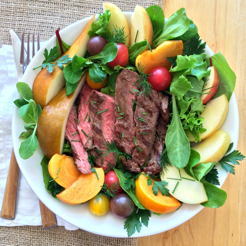Steak salad with fruits in a white bowl with fork on the left, on a burlap and wood background