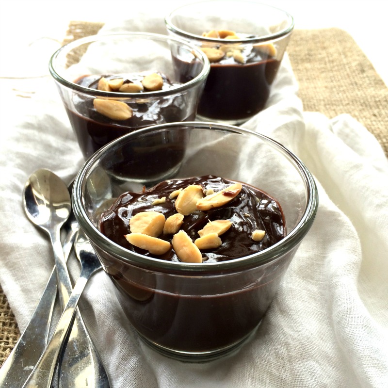 3 glass cups of dark chocolate pudding with nuts on top, sitting on fabric with spoons alongside