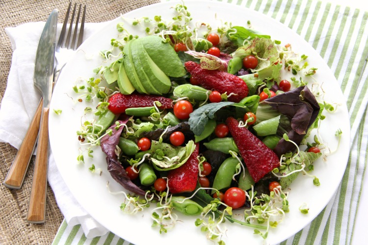 Salad with many ingredients on a white plate with fork and knife on left side, and green and white striped towel underneath.com