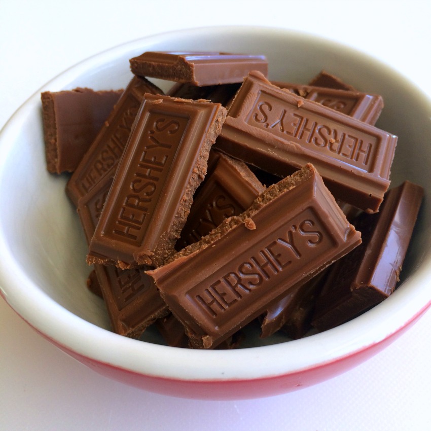 Broken Hershey Bars in a small white bowl with a red exterior on a white counter top. 
