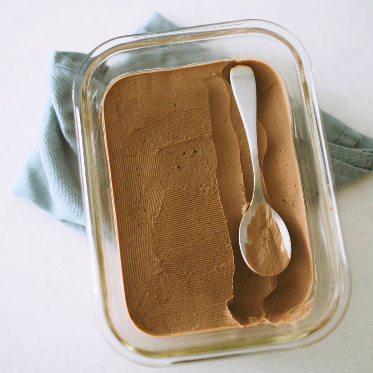 A silver spoon sits atop a rectangular glass container of milk chocolate ice cream, with a blue tea towel underneath. 