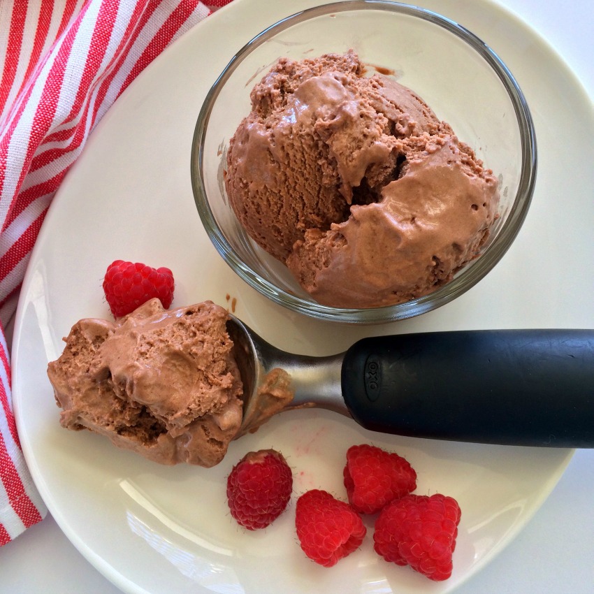 Black-handled scooper of No-Churn Fluffy Milk Chocolate Ice Cream rests on a white plate with raspberries alongside, and another scoop in a clear glass bowl. A red striped tea towel is on the left. 