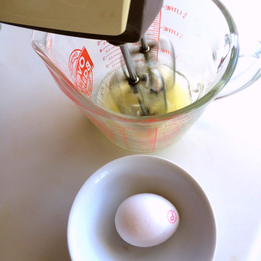 Hand mixer whips egg whites in a glass measuring cup. One white egg sits in a white bowl in the foreground. 