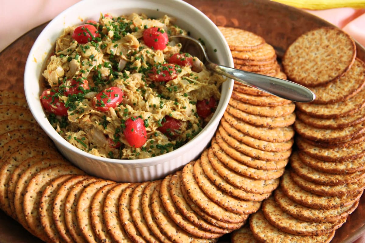 Coronation Turkey Salad dotted with cherry tomatoes on top in a white bowl set on a tray surrounded by crackers, with a spoon in the bowl