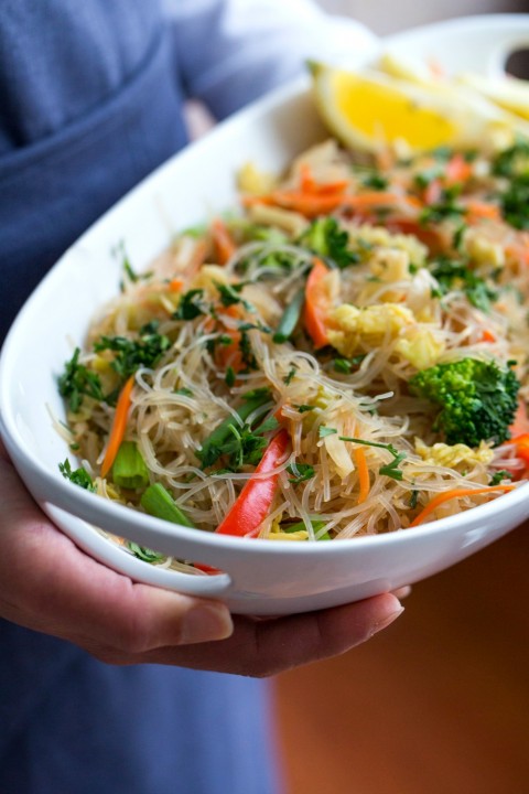 Stir-Fried Cellophane Noodles (vegan pancit) in a white bowl, held by a person in a blue apron