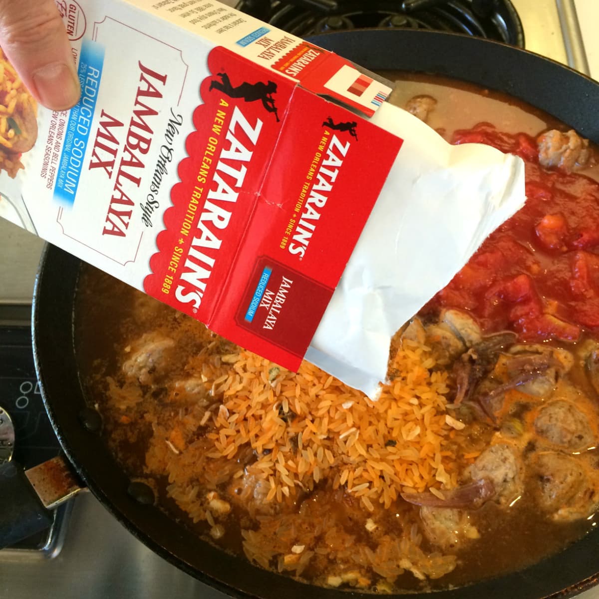 A hand pours orange colored rice mix into a skillet with sausage, water and tomatoes