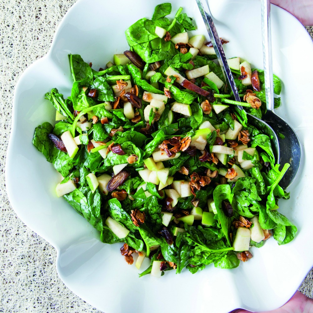 Green leafy Charoset Salad with apples, dates and nuts in a white bowl with silver serving spoons in the bowl