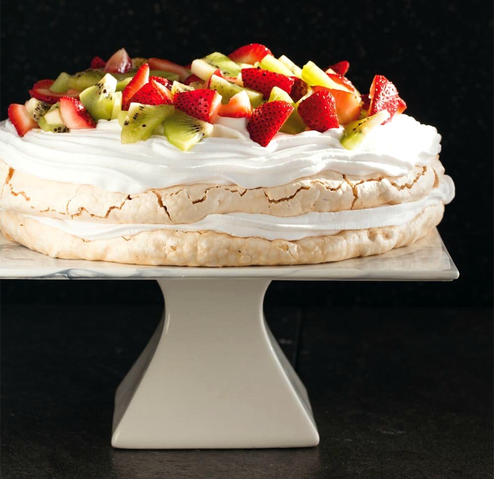 2 layer Pavlova dessert with white whipped cream and strawberries and kiwi on top on a white cake stand set against a black background