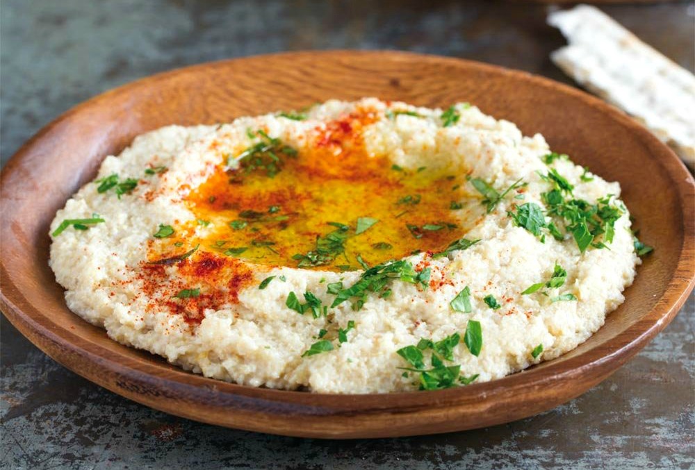 White hummus with green parsley and red paprika on top in a wooden bowl