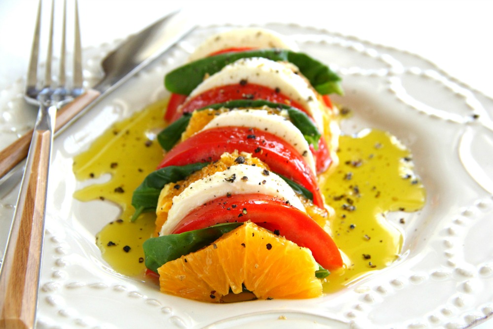 Alternating layers of orange, tomato, Mozzarella cheese and basil on a white plate with a form alongside and olive oil over the top. 