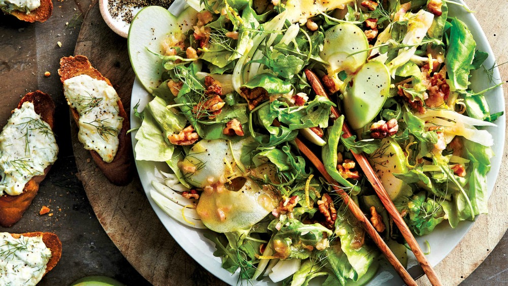 leafy arugula apple fennel salad on a white plate with wooden tongs and slices of bread alongside 