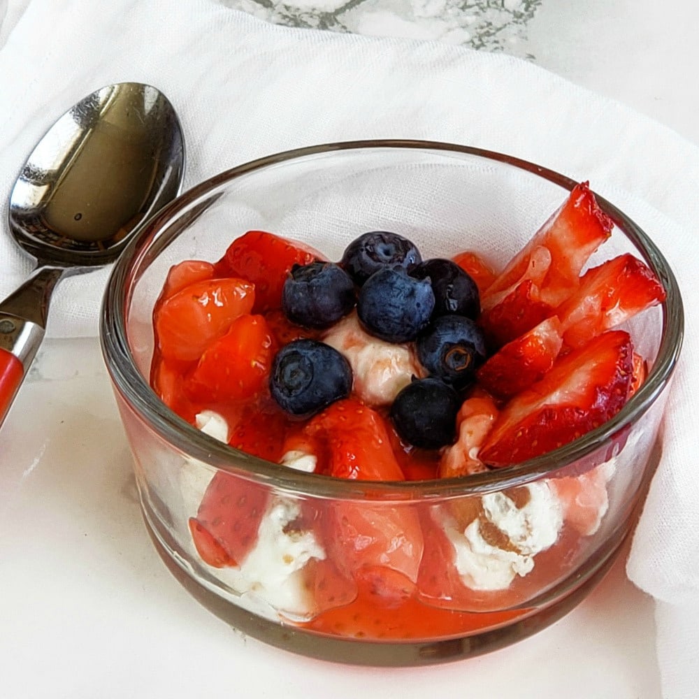 Strawberries, blueberries, cake cubes and whipped cream in a glass bowl with a spoon alongside on a marble counterom