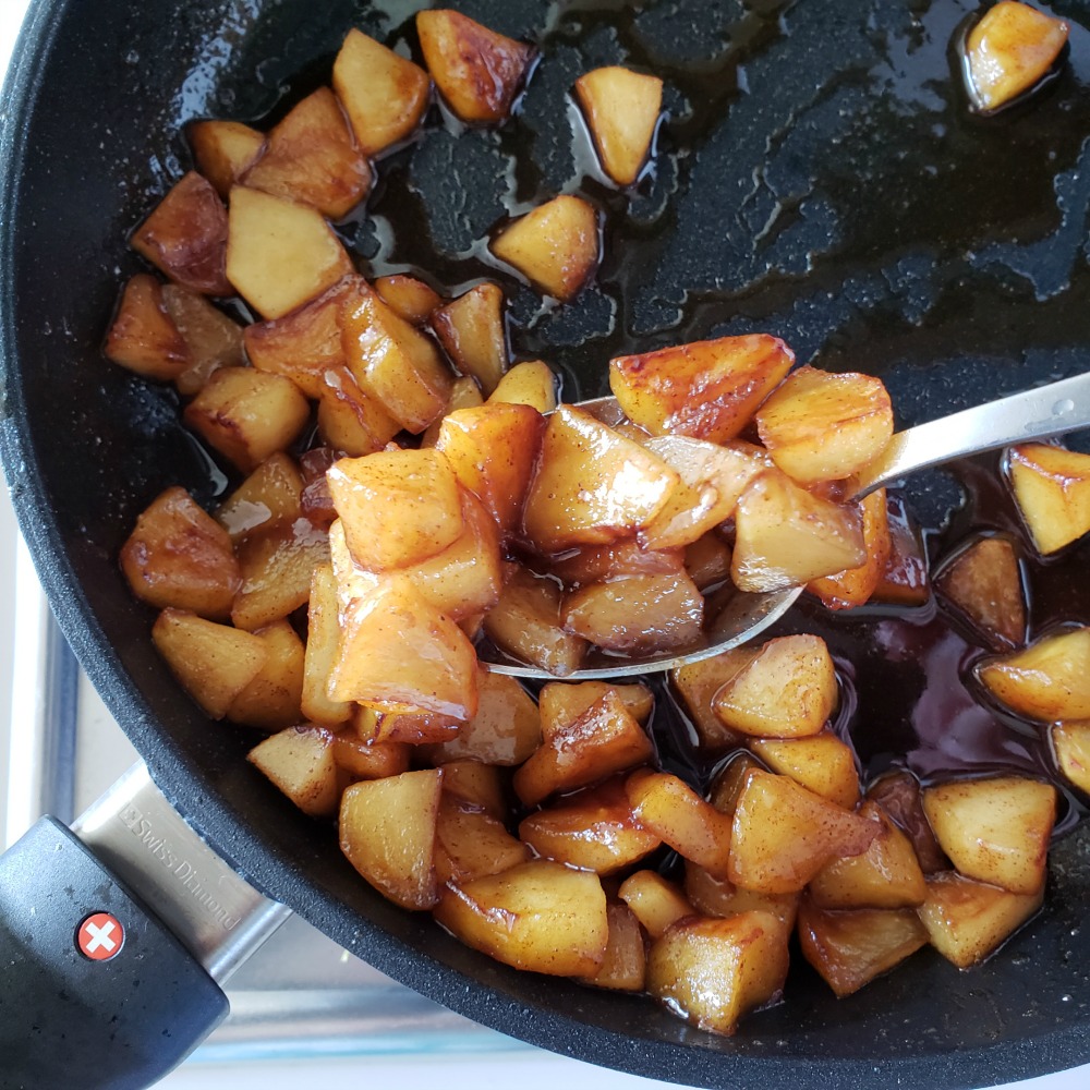 Spoon lifting caramelized apples in cinnamon maple syrup in the black skillet