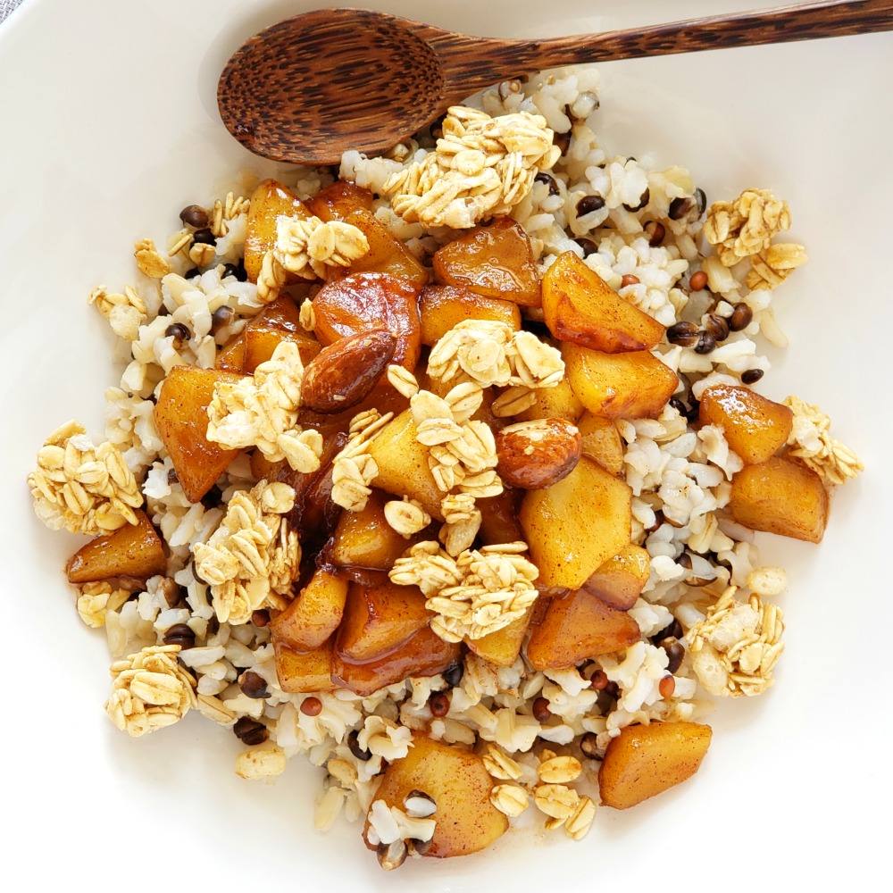 Apples grains and granola in a white bowl with a wooden spoon in the bowl