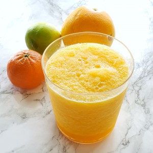 Sore Throat Slushy in a clear glass with a tangerine, orange and lime behind the glass on a white marble counter